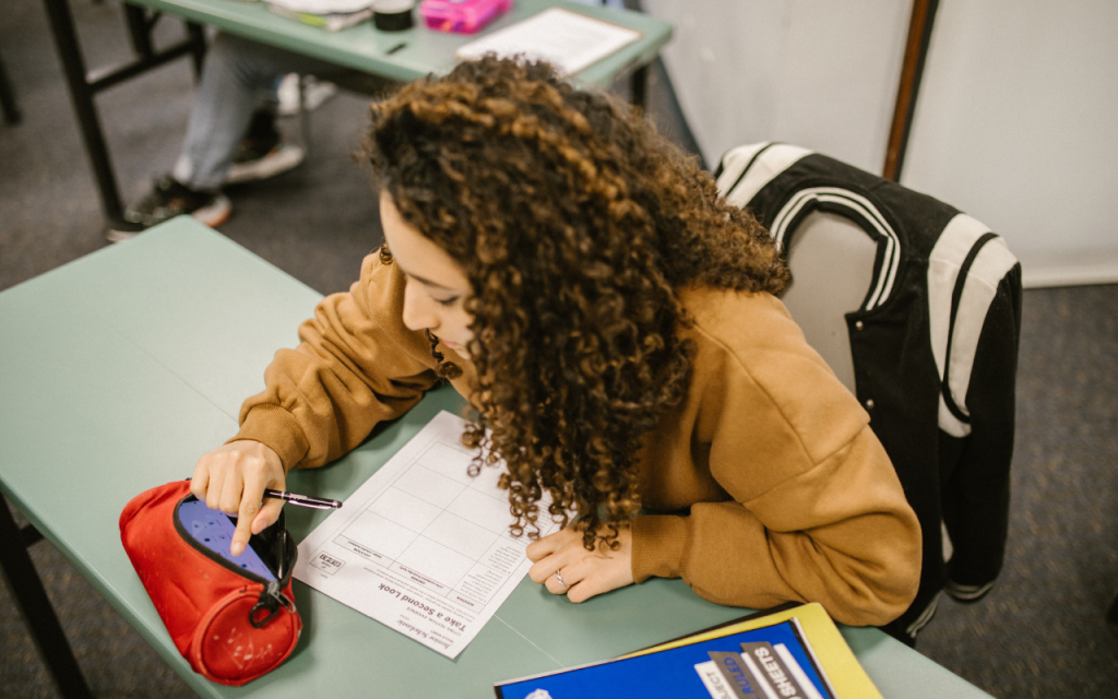 undergraduate student in brown sweatshirt sits at desk looking through pencil case