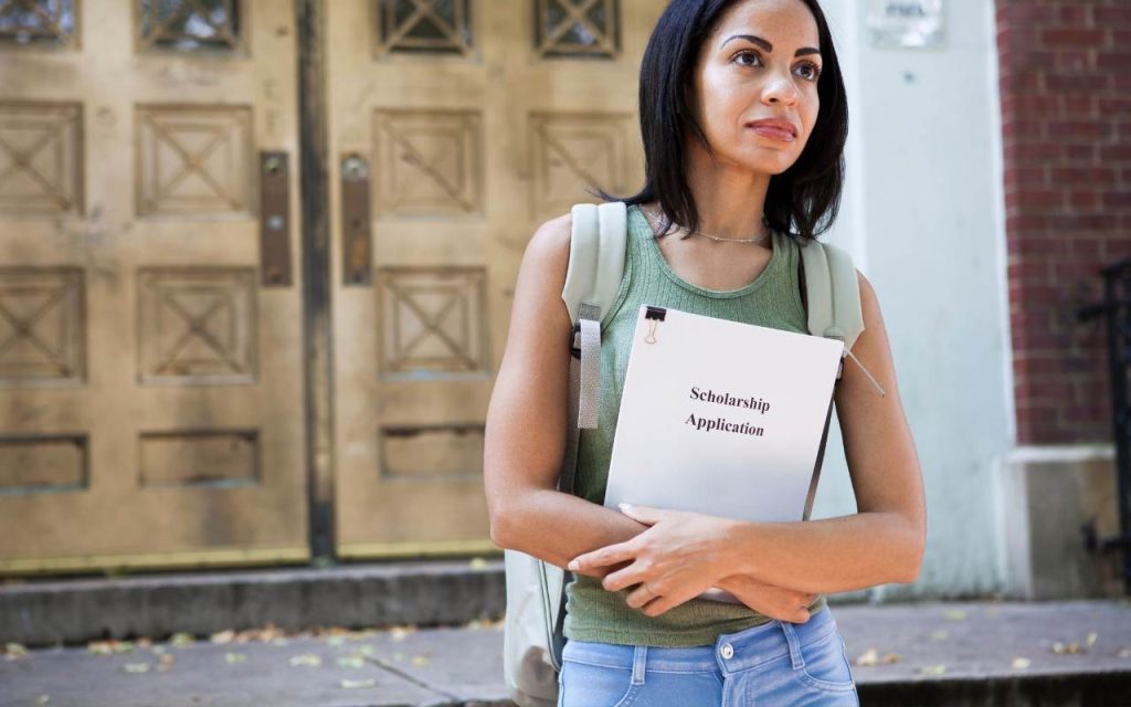 woman with backpack carries stack of paper labeled 'scholarship application'