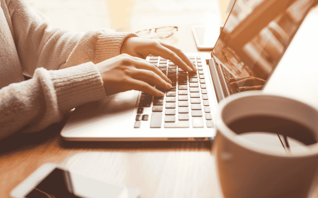 person in sweater types on laptop with mug of coffee