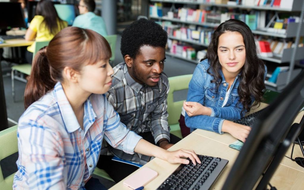 Three students sit in front of desktop computer in college library