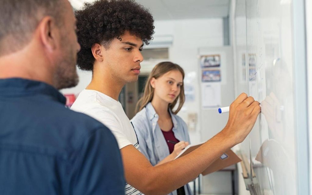 two students write on whiteboard while professor observes