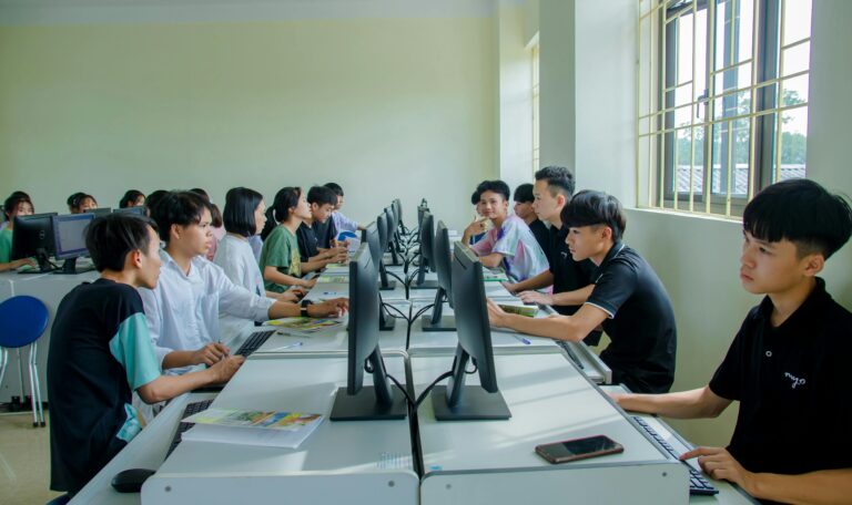 A group of young students working on desktop computers in a bright, modern computer lab or classroom setting. The students are seated in rows facing each other at white desks, with some engaged in discussion while others focus on their monitors. Natural light streams in from a large window on the right.