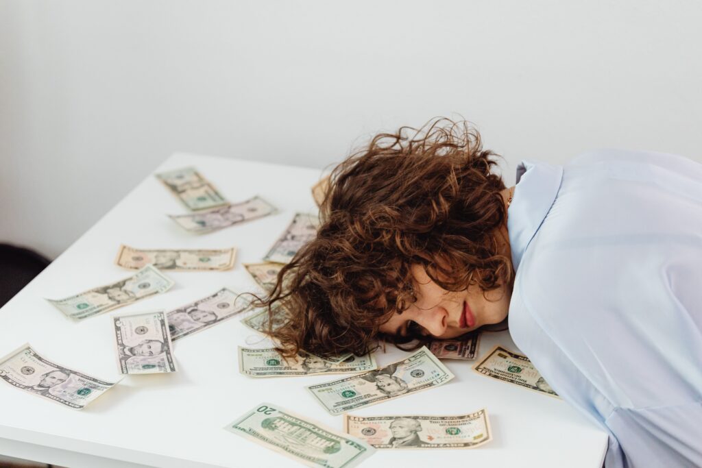 A woman with curly hair resting her head on a table covered in US dollar bills